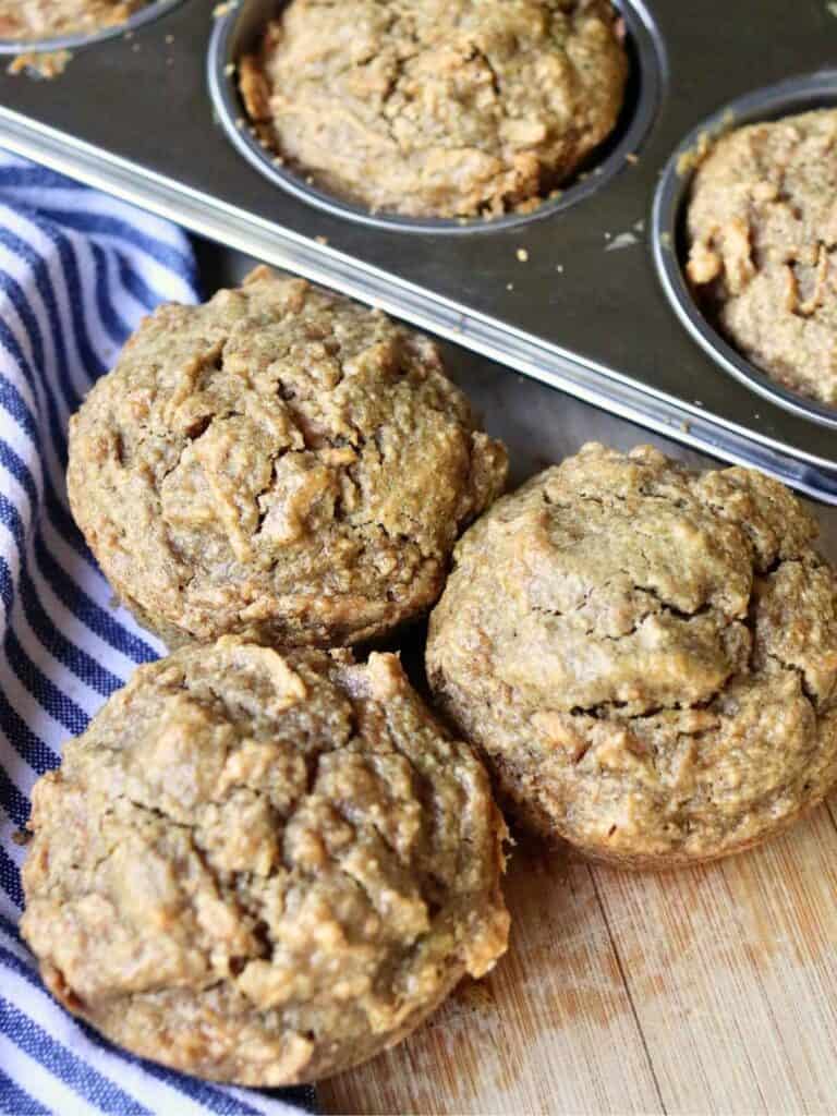 3 freshly baked apple oat muffins in a muffin tin and on a wooden surface, next to a striped cloth napkin.