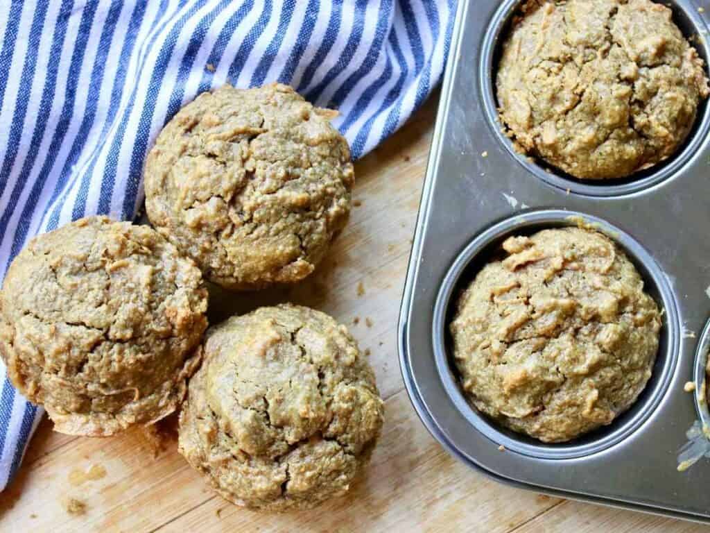Freshly baked apple oat muffins in a muffin tin and on a wooden surface, next to a striped kitchen towel.