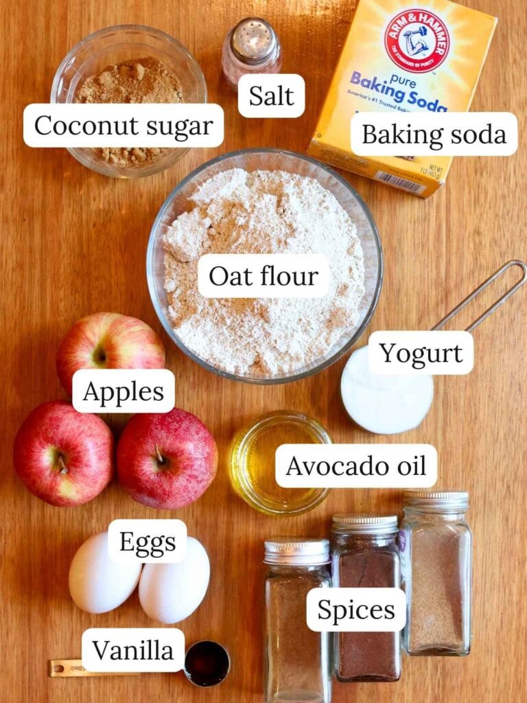 Flat lay of healthy apple oat muffin ingredients including oat flour, apples, eggs, coconut sugar, yogurt, avocado oil, vanilla, baking soda, salt, and spices on a wooden surface.