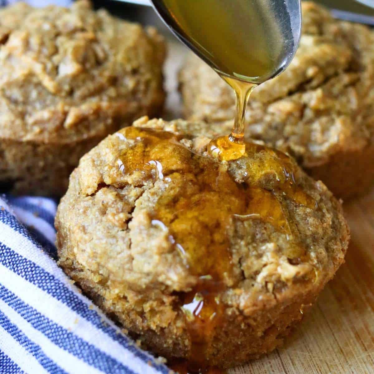Close-up view of freshly baked apple oat muffin being drizzled with honey, highlighting moist texture and wholesome ingredients with more muffins in the background.
