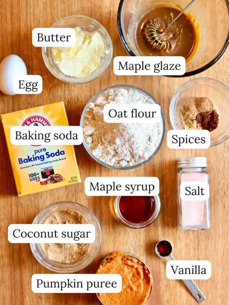 Ingredients for gluten-free pumpkin cookies are arranged on a wooden surface, labeled: butter, egg, baking soda, oat flour, coconut sugar, pumpkin puree, maple glaze, spices, maple syrup, salt, and vanilla. A warm, cozy preparation mood is conveyed.