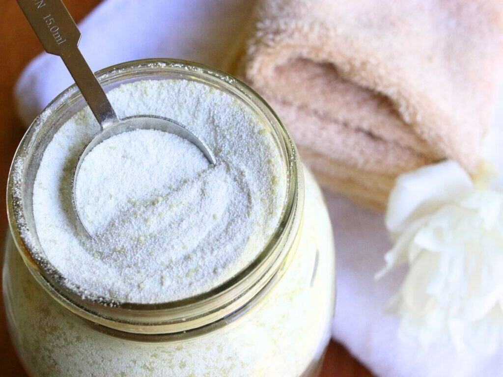 Closeup of a tablespoon in a glass jar full of homemade laundry powder beside a stack of clean towels.