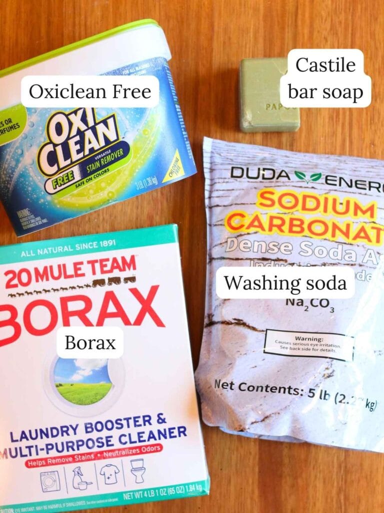 Flat lay of laundry powder ingredients including Oxiclean Free, Castile bar soap, borax, and washing soda on a wooden surface.