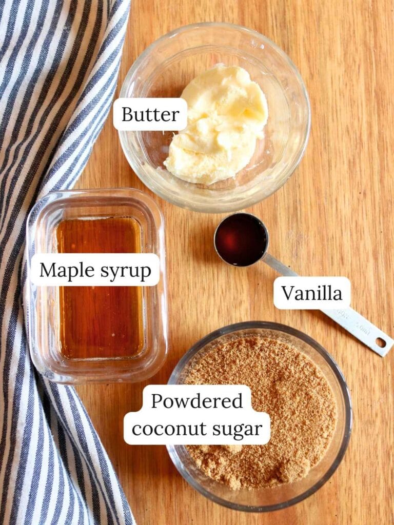 Ingredients for maple glaze on a wooden table: butter in a bowl, maple syrup in a glass dish, powdered coconut sugar in another bowl, and vanilla in a spoon with a striped cloth nearby.