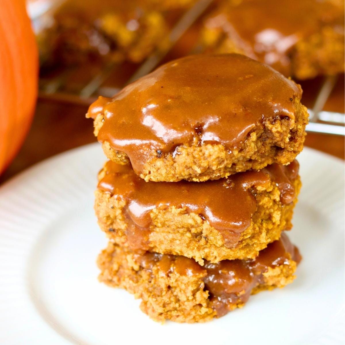 Stack of three gluten free pumpkin cookies dripping with rich maple glaze on a white plate. A warm, cozy autumn treat.