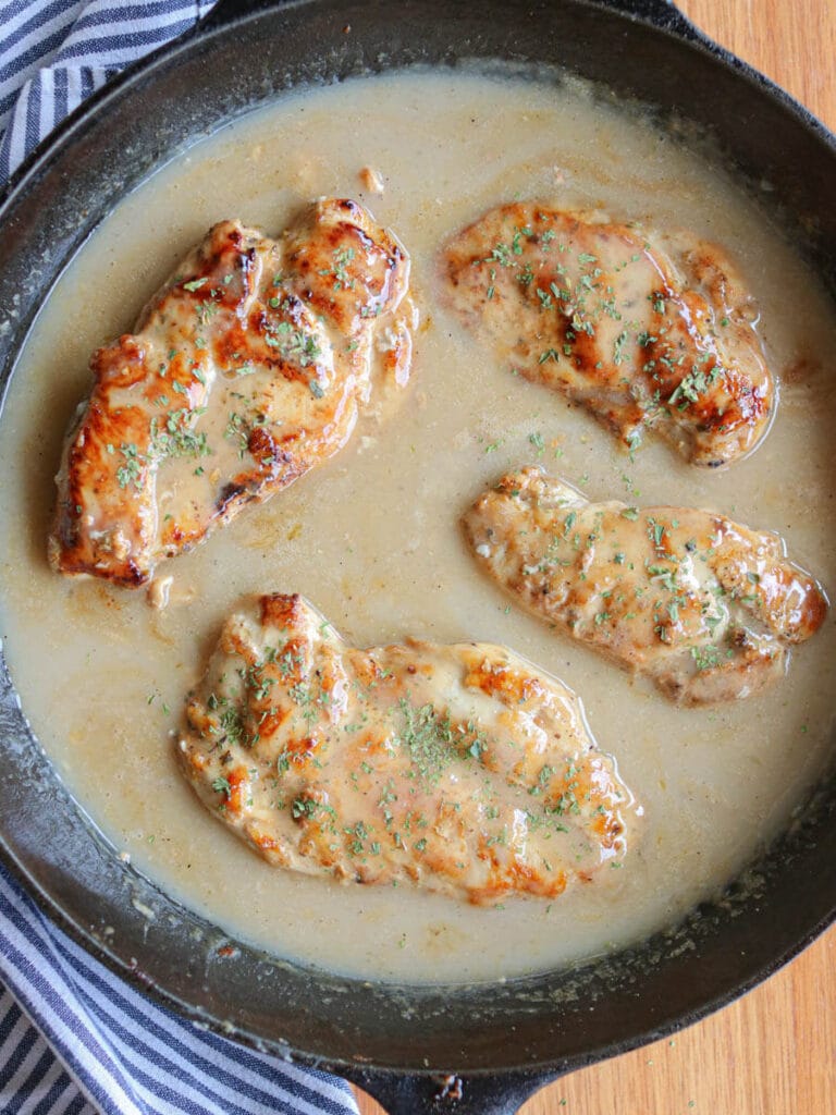 Four seasoned chicken breasts smothered in gravy in a cast iron skillet beside a striped cloth napkin.