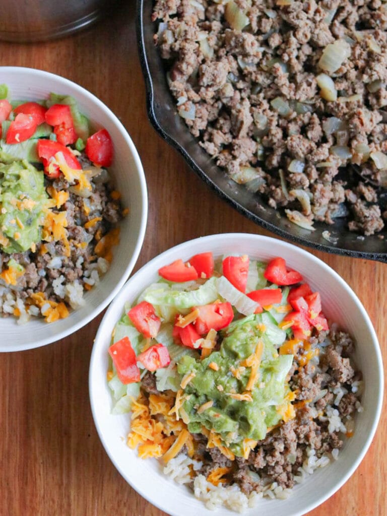 2 bowls with ground beef and rice topped with shredded cheese, guacamole, and chopped tomatoes on a wooden table with a cast iron skillet of ground beef in the background.