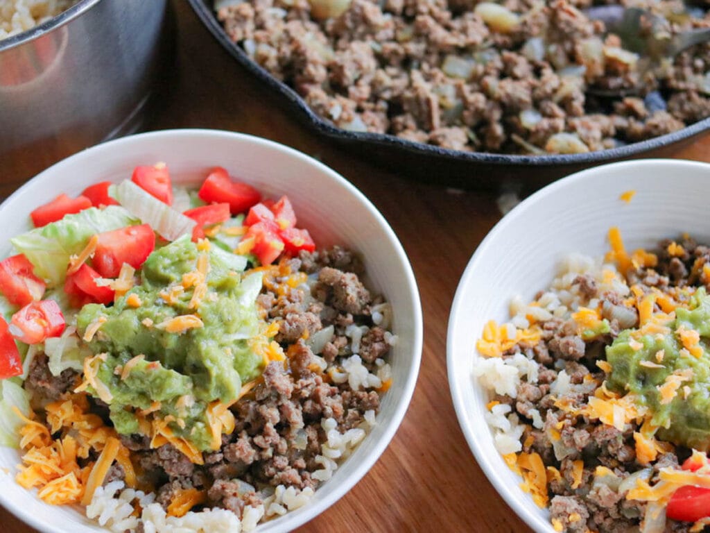 2 bowls with ground beef and rice topped with shredded cheese, guacamole, and chopped tomatoes with a skillet of ground beef in the background.
