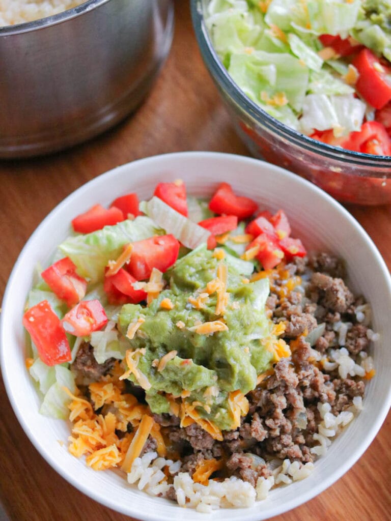 Bowl full of ground beef and rice topped with shredded cheese, guacamole, and chopped tomatoes.