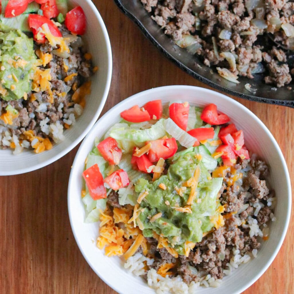 2 bowls with ground beef and rice topped with shredded cheese, guacamole, and chopped tomatoes.