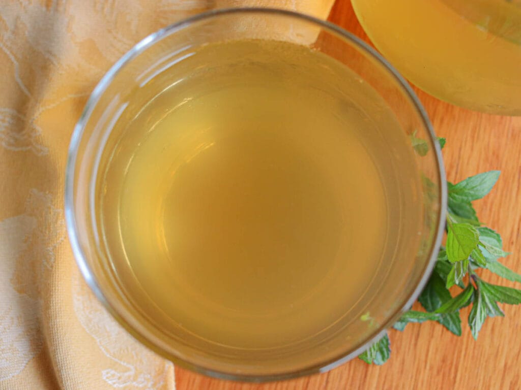 A top-down view of a glass of light yellow peppermint tea on a wooden surface. Peppermint leaves rest beside the glass, next to a beige cloth.