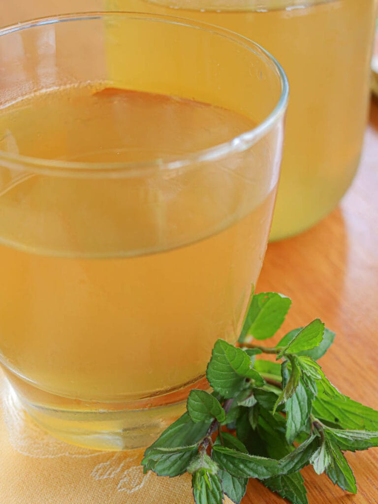 Clear glass of golden peppermint tea with fresh peppermint sprigs on a wooden table. The setting is warm, inviting, and soothing.