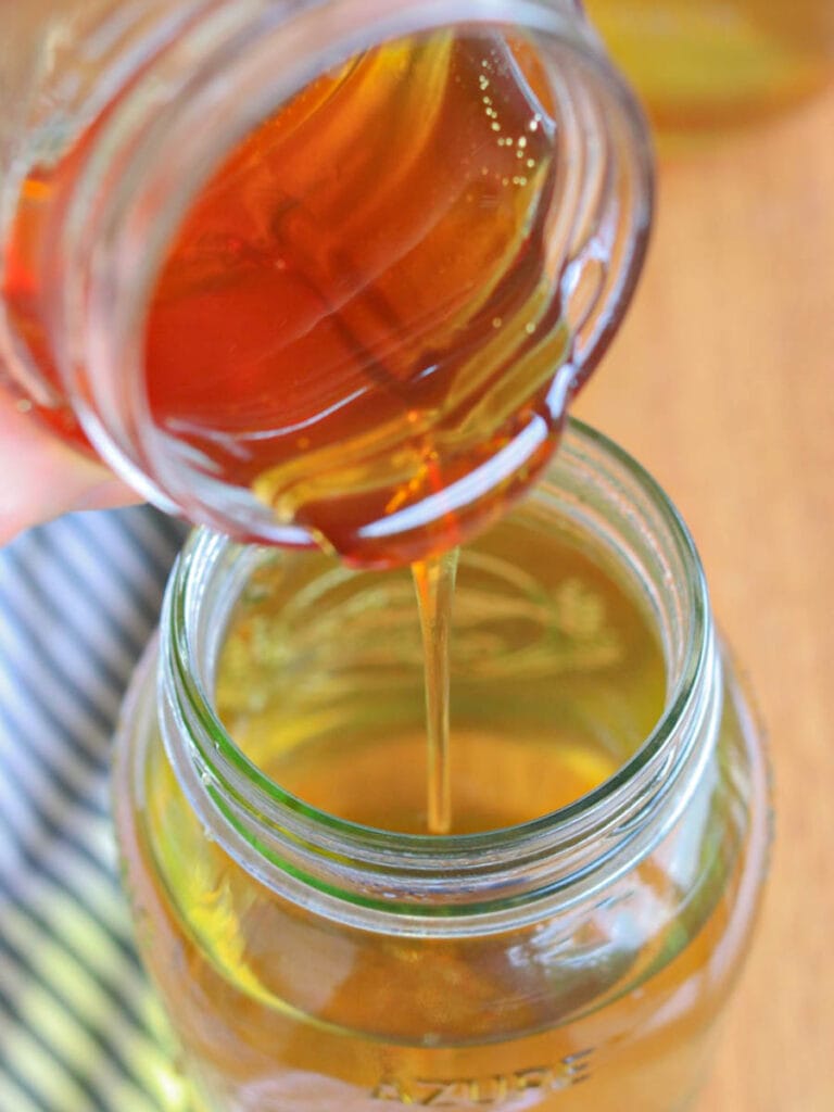 Pouring golden honey from a small jar into a larger jar filled with peppermint tea. The honey's rich texture and amber color are illuminated under natural light, evoking sweetness.