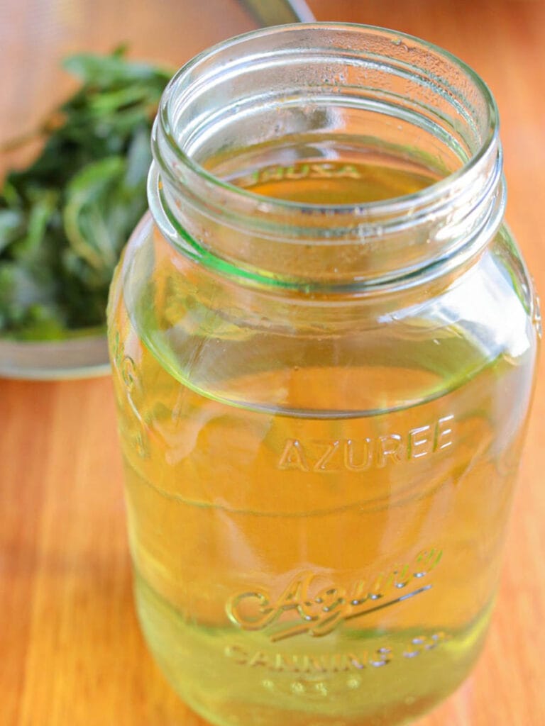 A glass jar filled with golden peppermint tea against a wooden table background. In the blurred background, fresh green peppermint leaves are visible in a metal strainer.