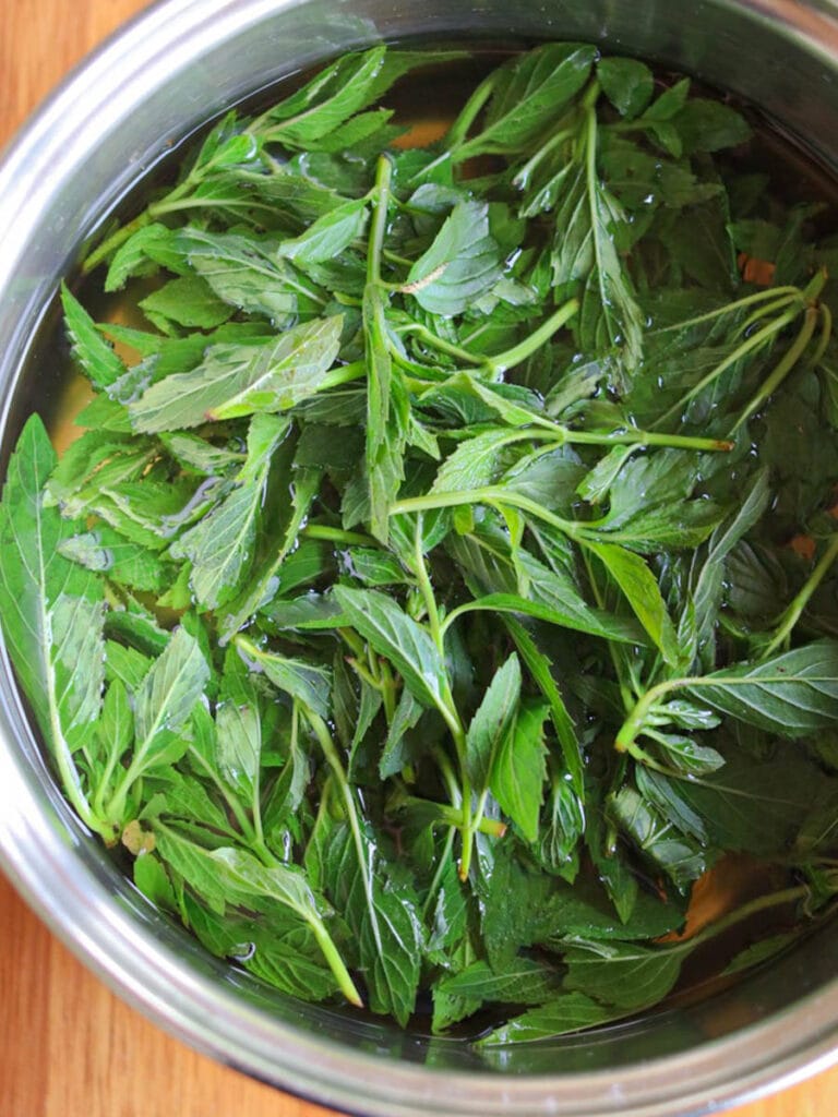 Fresh peppermint leaves submerged in water inside a shiny metal pot on a wooden surface, conveying a refreshing and natural tone.