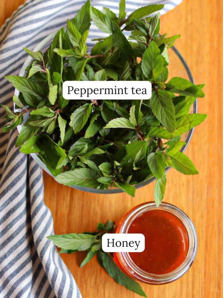 A bowl of fresh peppermint leaves beside a jar of honey on a wooden table, with a striped cloth nearby. Labels read "Peppermint tea" and "Honey".