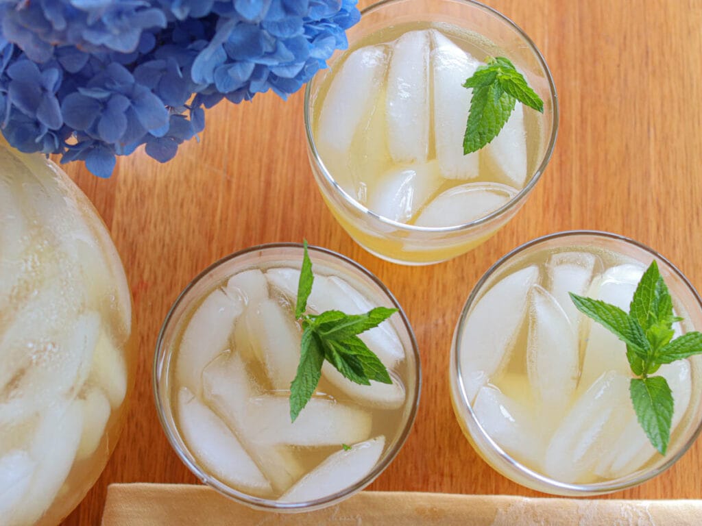 Top view of three glasses of iced spearmint tea with spearmint leaves on a wooden table, next to blue hydrangeas and a large pitcher, creating a refreshing summer vibe.