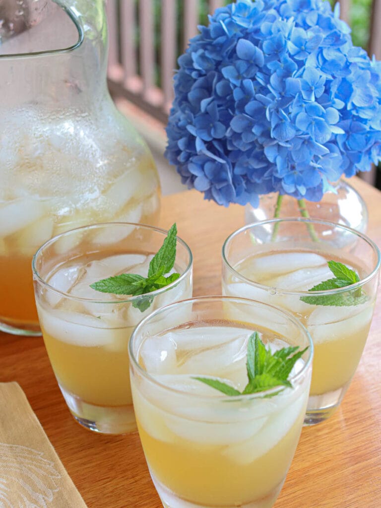 Three glasses of iced spearmint tea with spearmint garnish sit on a wooden table beside a pitcher and a vibrant blue hydrangea, evoking a relaxing summer vibe.
