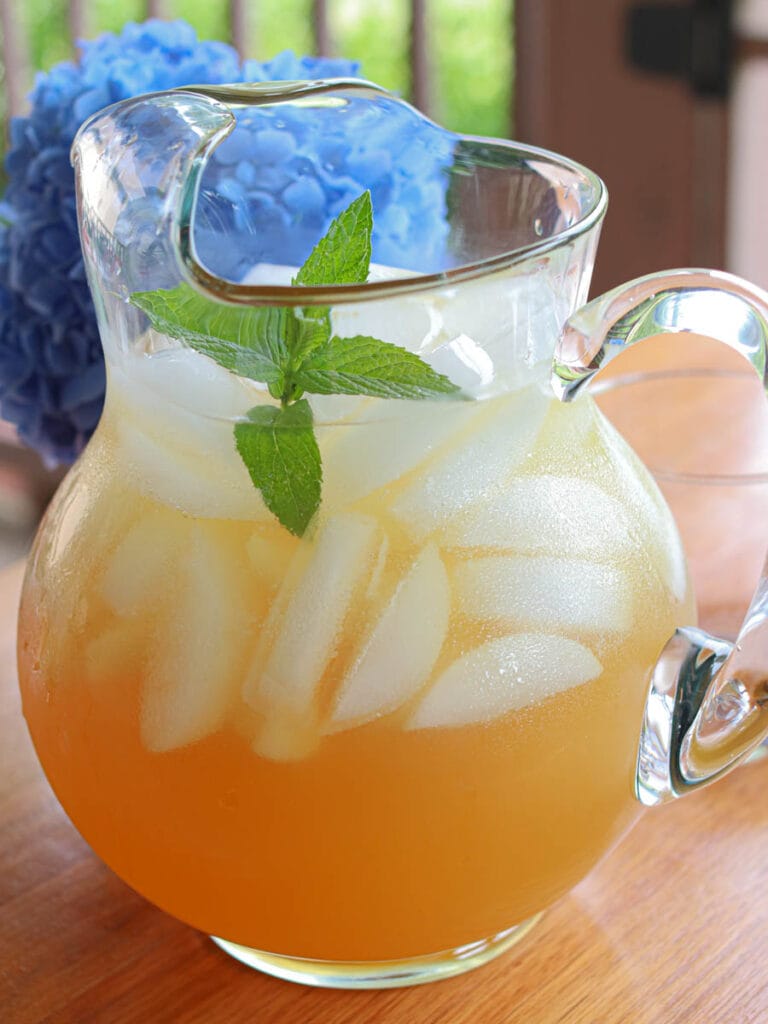 A glass pitcher of iced spearmint tea with spearmint leaves and ice cubes, on a wooden table. A blue hydrangea flower is in the background, giving a refreshing summer vibe.