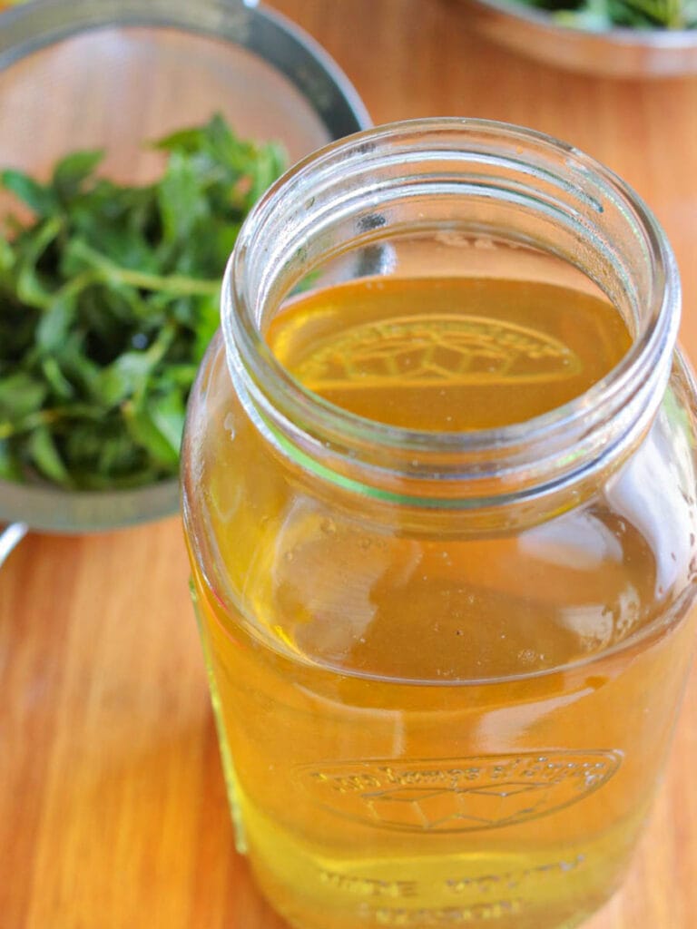A glass jar filled with golden spearmint tea against a wooden table background. In the blurred background, fresh green tea leaves are visible in a metal strainer.