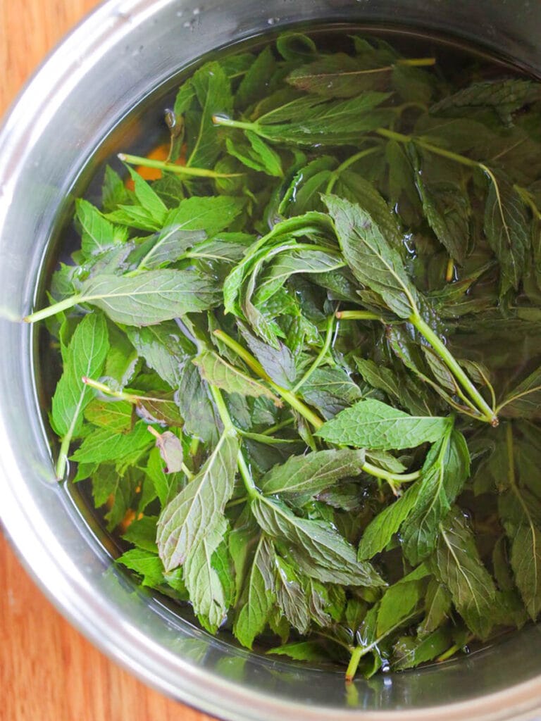 Fresh spearmint leaves submerged in water inside a shiny metal pot on a wooden surface, conveying a refreshing and natural tone.