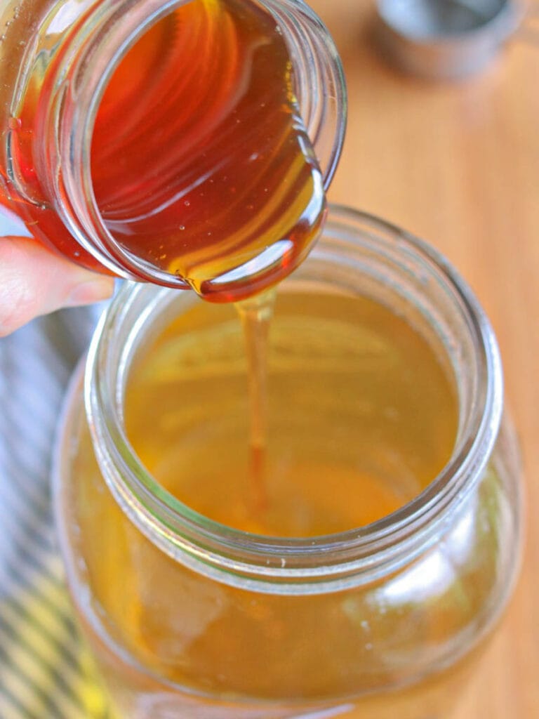Pouring golden honey from a small jar into a larger jar full of spearmint tea. The honey's rich texture and amber color are illuminated under natural light, evoking sweetness.