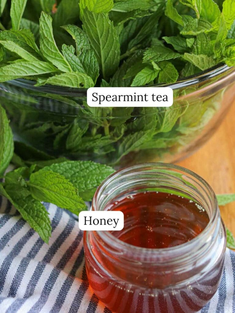 A bowl of fresh spearmint leaves beside a jar of honey on a wooden table, with a striped cloth nearby. Labels read "Spearmint tea" and "Honey".