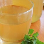 Glass of golden peppermint tea with fresh peppermint sprigs on a wooden surface. The setting is warm, inviting, and soothing.