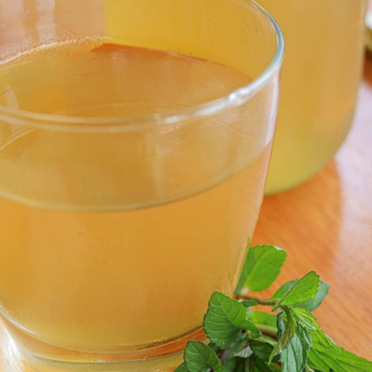 Glass of golden peppermint tea with fresh peppermint sprigs on a wooden surface. The setting is warm, inviting, and soothing.