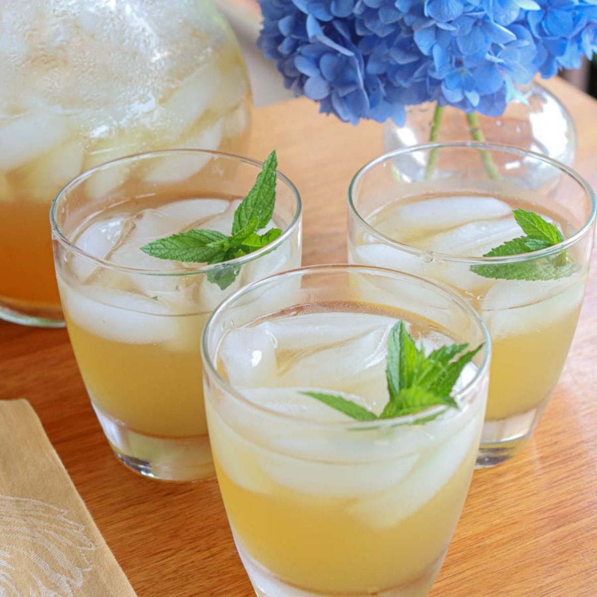 Closeup of three glasses of iced spearmint tea with spearmint garnish sit on a wooden table beside a pitcher and a vibrant blue hydrangea, evoking a relaxing summer vibe.