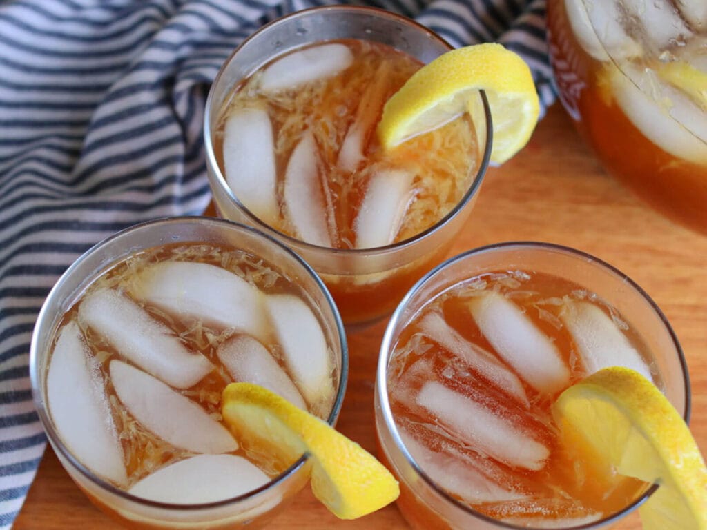 Three glasses of healthy lemonade with lemon wedges on a wooden table. Ice cubes float in the amber drink. A striped cloth adds a casual, refreshing vibe.