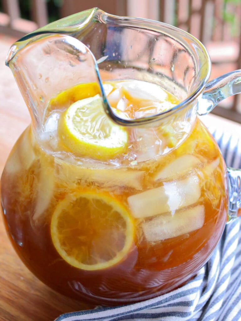 A glass pitcher filled with lemonade (healthy), featuring lemon slices and ice cubes. It's set on a wooden table with a striped cloth, creating a refreshing, summery vibe.