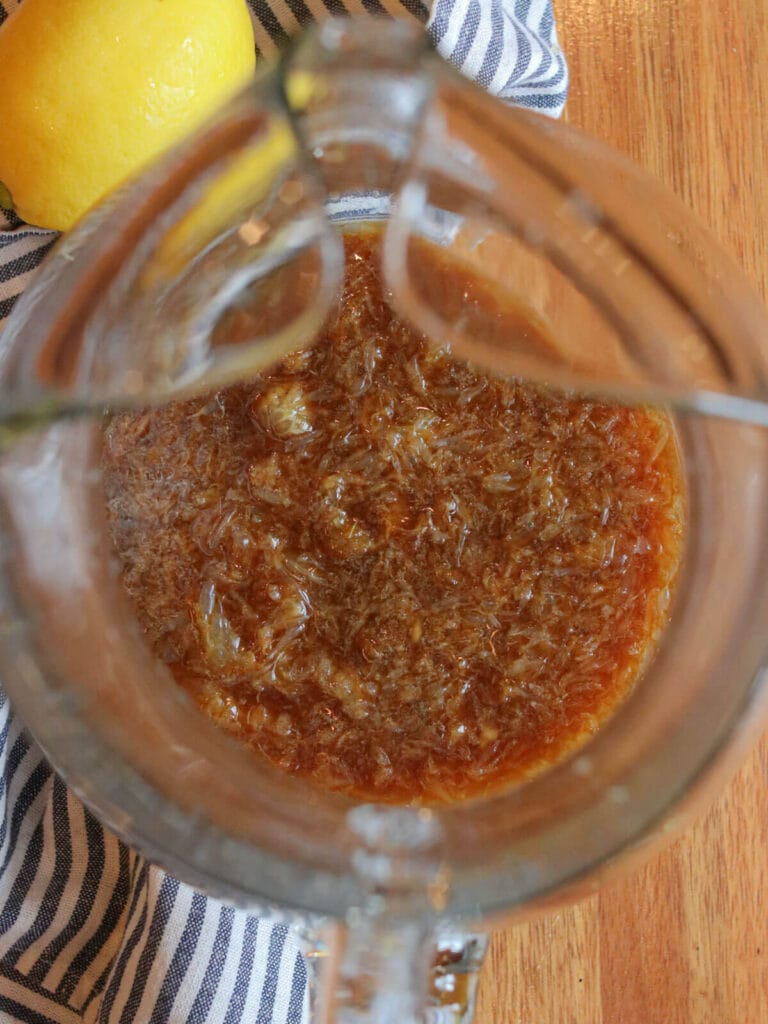 Top view of a glass pitcher filled with brown coconut sugar and lemon juice and pulp placed on a striped cloth. A fresh lemon is adjacent, suggesting a refreshing summer drink.