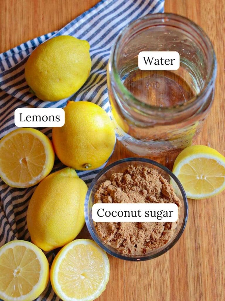 Jar of water, whole and halved lemons, and a bowl of coconut sugar on a striped cloth; evokes freshness and natural sweetness.