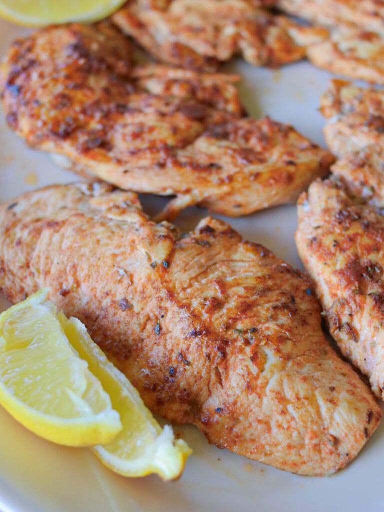 Closeup of a thin sliced chicken breast on a white serving plate with two fresh lemon slices on the side.