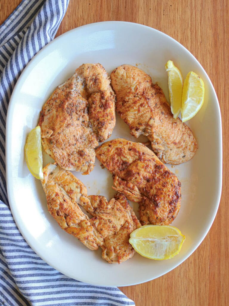 Overhead view of a white serving plate with thin cut chicken garnished with lemon slices on a wooden background.