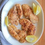 A white plate with four thin cut chicken breasts garnished with fresh lemon slices on a wooden table with a striped cloth napkin.