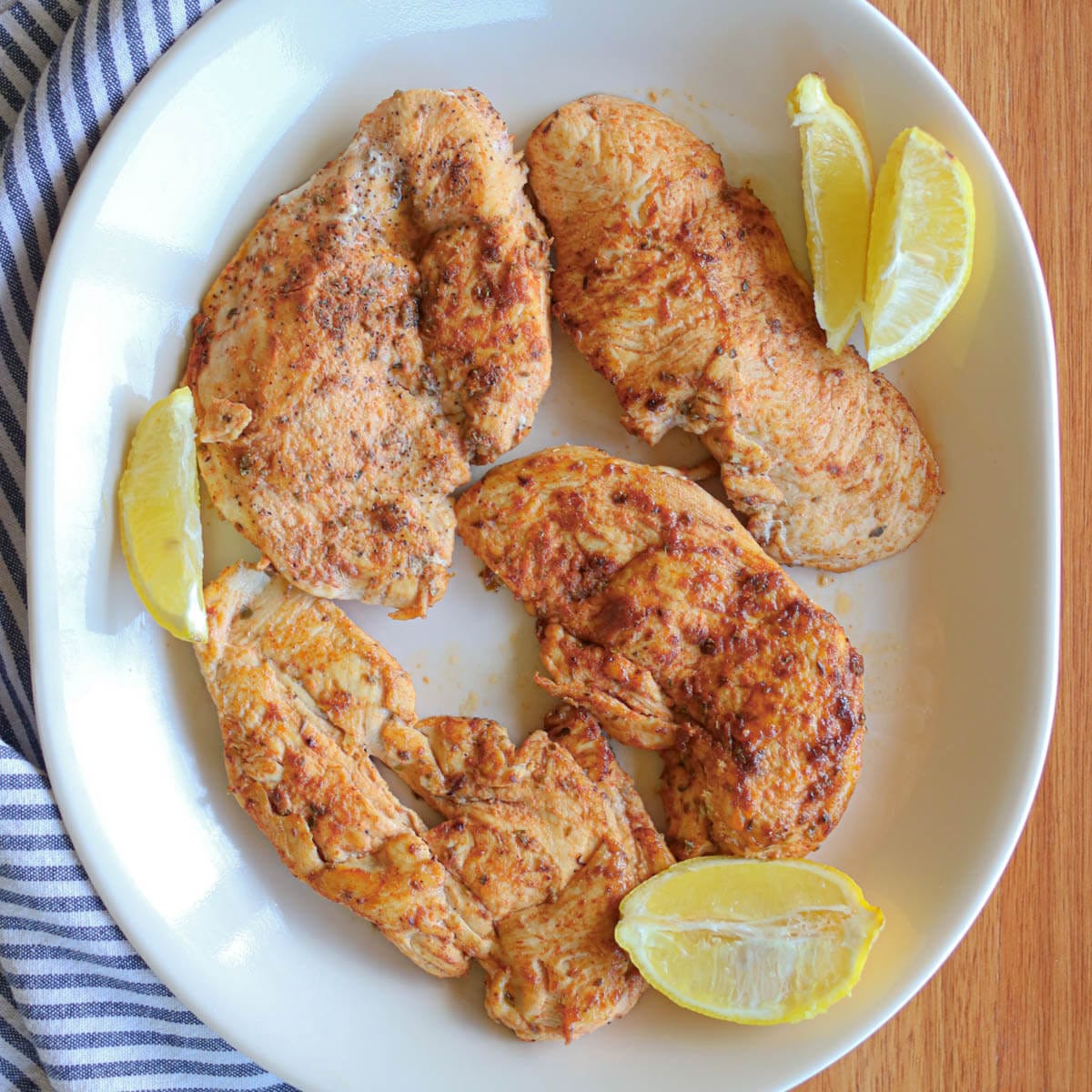 A white plate with four thin cut chicken breasts garnished with fresh lemon slices on a wooden table with a striped cloth napkin.