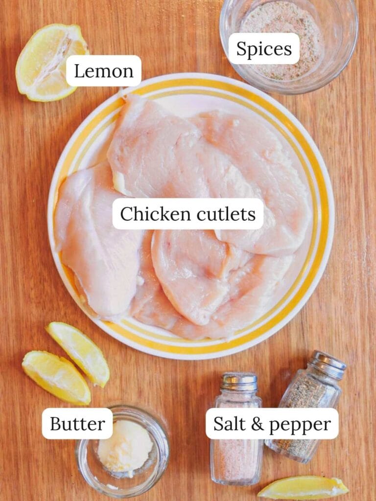 Labeled ingredients for thin cut chicken breast on a wooden table including lemon, spices, butter, salt, pepper, and chicken cutlets.