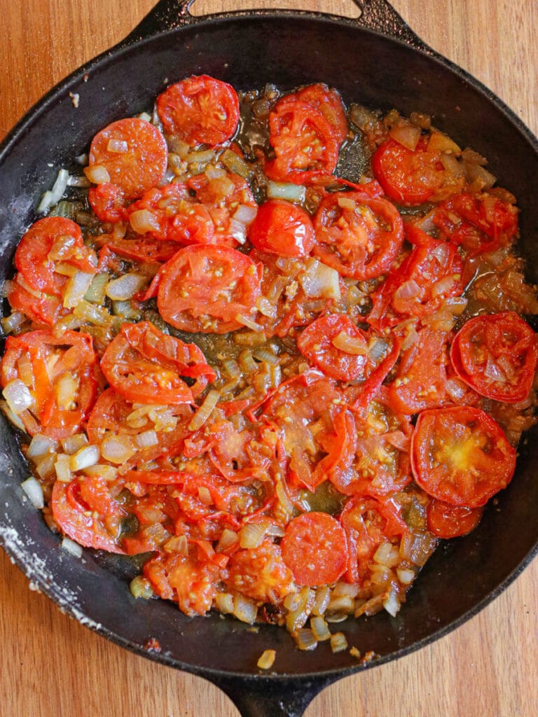 Tomato slices, chopped onion, and minced garlic cooking in a cast iron skillet.