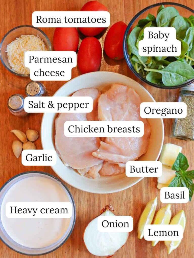 Labeled ingredients on a wooden background including Roma tomatoes, parmesan cheese, baby spinach, salt and pepper, oregano, chicken breasts, garlic, butter, basil, heavy cream, onion, and lemon slices.