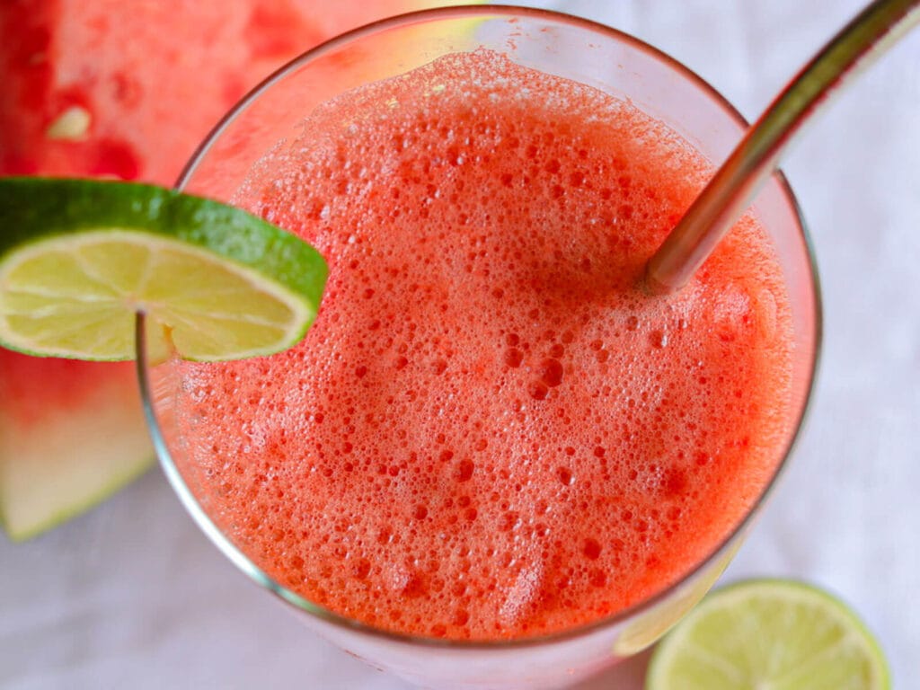 Close-up of frothy red watermelon juice in a glass with a metal straw, garnished with a lime slice. Bright and refreshing tone.