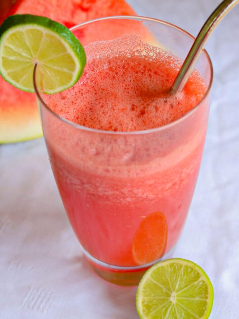 A cold glass of watermelon juice is topped with frothy bubbles and a lime slice on the rim, with a metal straw. Bright and summery vibe.