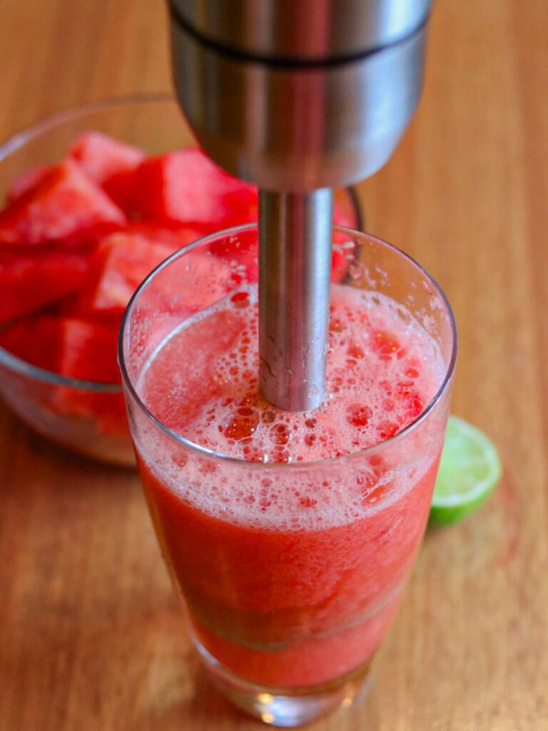 An immersion blender froths vibrant red watermelon juice in a glass on a wooden table. Behind it, a bowl of watermelon chunks and a lime wedge are visible.