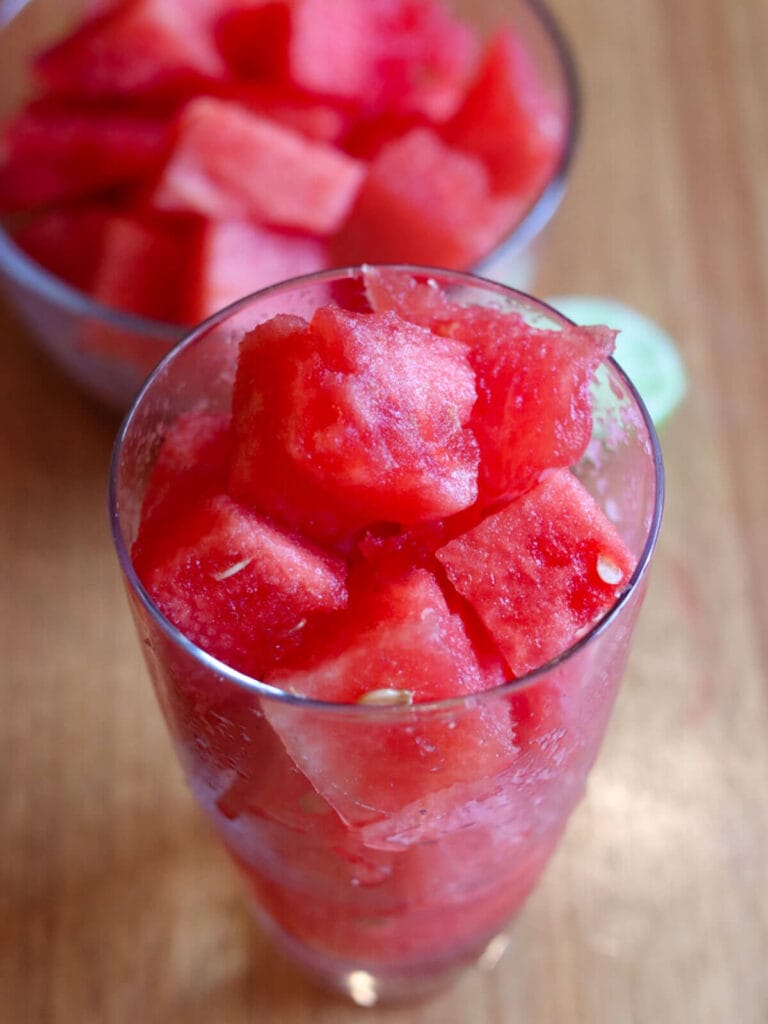 Glass filled with vibrant red watermelon cubes, lime juice, and coconut water on a wooden table, conveying a fresh and summery feel. A bowl with more pieces is in the background.