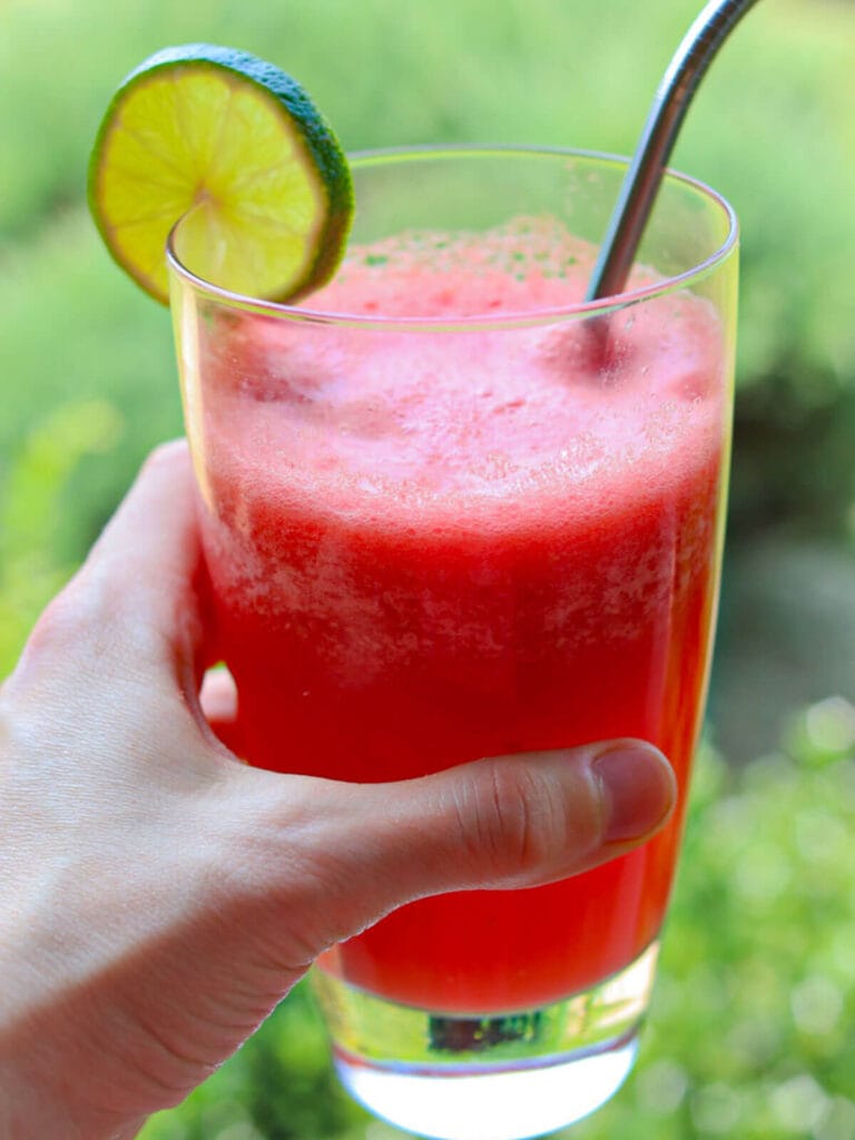 A hand holds refreshing red watermelon juice in a clear glass, garnished with a lime slice and a metal straw, set against a blurry green background.