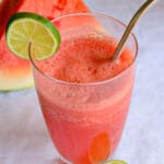 A refreshing glass of watermelon juice is topped with frothy bubbles and a lime slice on the rim, with a metal straw. Bright and summery vibe.