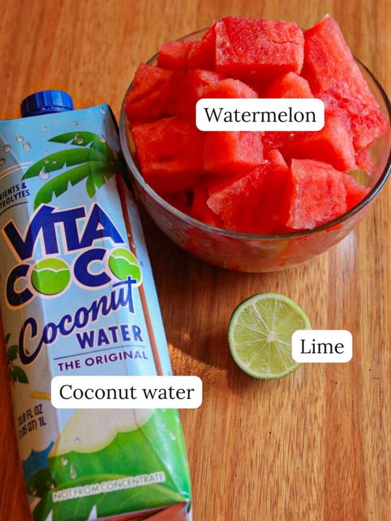A bowl of vibrant red watermelon cubes beside a blue carton of coconut water and a halved lime on a wooden surface. Refreshing and tropical.