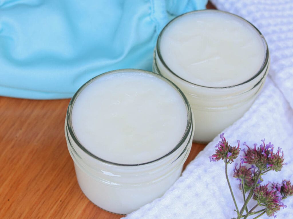 Two glass jars of baby balm on a wooden table with a white towel and fresh purple flowers.