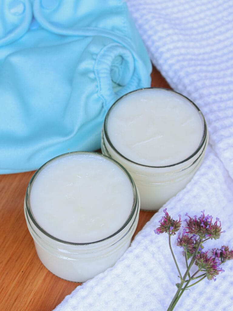 Two glass jars of baby balm on a wooden surface with a white towel and blue cloth diaper.